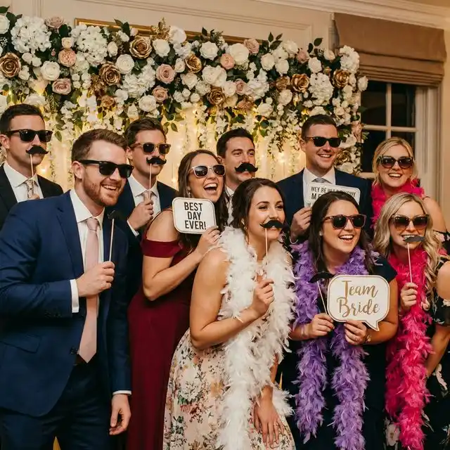 Invitados divirtiéndose en un photobooth de boda con accesorios divertidos y fondo decorado