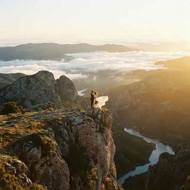 Pareja silueteada en un acantilado de montaña al atardecer con el vestido de novia al viento, paisaje épico